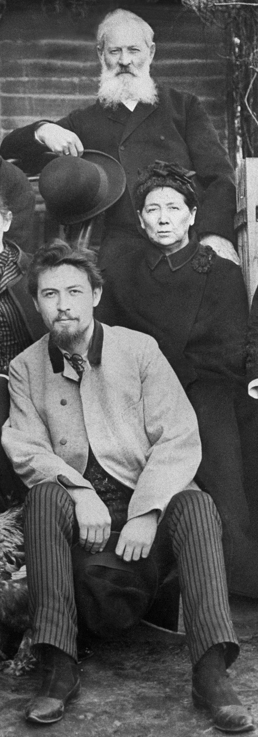 Black and white photograph of the Chekhov family and friends in 1890, showing Anton Chekhov seated in the front row center wearing a light-colored jacket, surrounded by his siblings and family members posed on outdoor steps